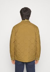 Gesteppte, olivgrüne Jacke mit matter Oberfläche, mit abgerundetem Muster, Kragen und Druckknöpfen, getragen über blauen Jeanshosen.