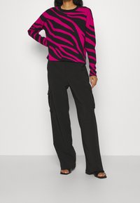 Maglione a righe zebra rosa e nero con maniche lunghe, abbinato a pantaloni cargo neri a vestibilità ampia e sandali neri con punta aperta.
