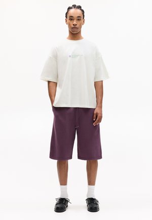 White oversized T-shirt with a small graphic, paired with knee-length purple shorts. Accessories include black shoes and white socks.