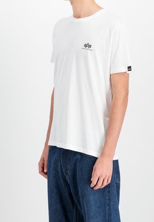 Personne portant un t-shirt blanc Alpha Industries et un jean bleu, debout de côté sur un fond clair uni.