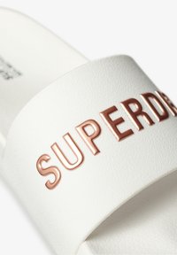 White slide sandal made of textured synthetic material featuring a wide strap embossed with the word "SUPERDRY" in rose gold lettering.