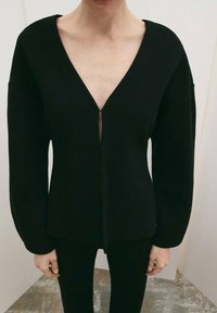 Čierny pletený top s hlbokým výstrihom do V a dlhými rukávmi. Má predné zapínanie a jemnú textúru, priliehavý v páse.