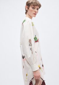 Φόρεμα από λινό σε εκρού χρώμα, υπερμεγέθες, με πολύχρωμη κεντητική με λουλούδια, μακριά μανίκια και ρεβέρ με πιέτες· μοντέλο κρατά μια μπορντό τσάντα.