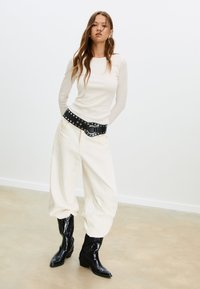 Top aderente a maniche lunghe color crema abbinato a pantaloni oversize color crema. Cintura nera con borchie e stivali da cowboy neri con punta affilata.