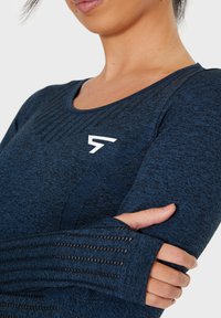 Dunkelblaues langärmliges Sportoberteil aus strukturiertem Stoff, mit rundem Halsausschnitt und einem weißen Logo auf der Brust; verfügt über Daumenlöcher in den Bündchen.
