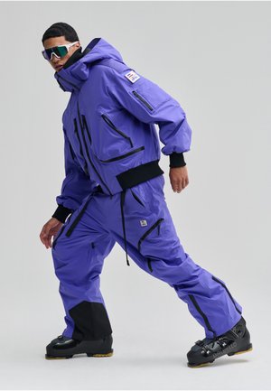 Lilla skijakke og bukser med sorte detaljer, udstyret med flere lynlåslommer og en stor hætte. Sorte skistøvler fuldender outfittet.