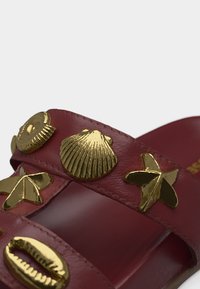 Κόκκινο σανδάλι με χρυσά διακοσμητικά εμπνευσμένα από τη θάλασσα, συμπεριλαμβανομένου ενός κοχυλιού, αστερία και σχημάτων κοχυλιών σε δερμάτινους ιμάντες.