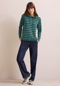 Felpa a righe verde e blu navy con cappuccio e cordino, abbinata a jeans blu scuro e sneakers multicolori. Vestibilità rilassata, design casual.