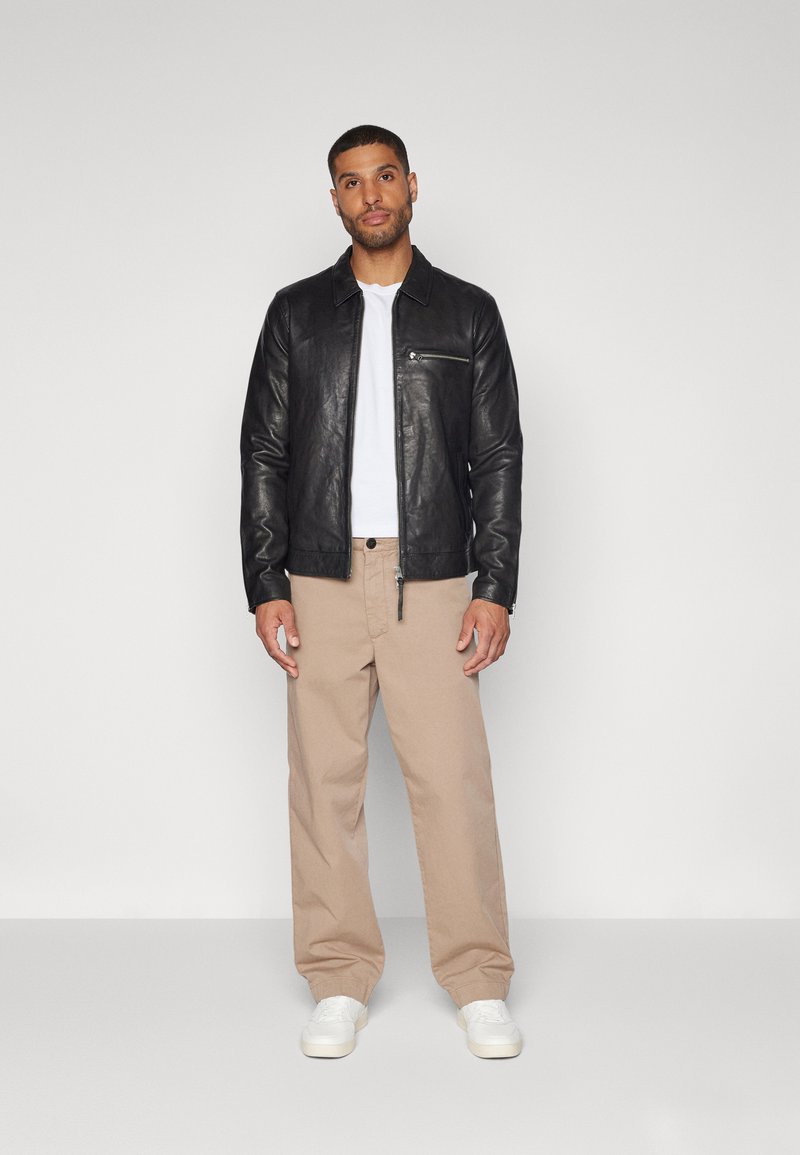 Schwarze Lederjacke mit einer Reißverschlusstasche, getragen über einem weißen Hemd. Kombiniert mit beigen Hosen und weißen Sneakern. Glatte Textur, taillierter Schnitt.