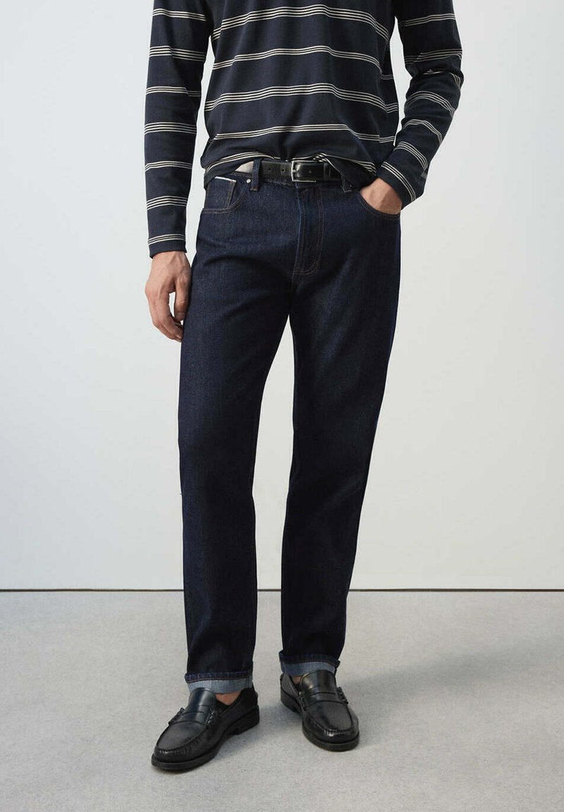 Homme portant un jean bleu foncé avec les revers roulés, une ceinture noire, des mocassins en cuir noir et une chemise bleu marine à manches longues avec des rayures blanches horizontales.