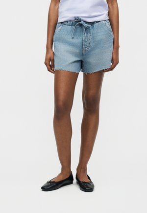 Isik, kes kannab helesiniseid teksast nööriga lühikesi pükse ja musti baleriinikingi, seisab valgel taustal.