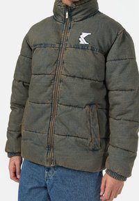 Puffjacka i mörkgrön tyg med quiltad struktur, hög krage, framdragkedja och broderad logotyp på bröstet. Har sidofickor.