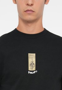 Juodas medvilninis marškinėliai su auksiniu „MARVEL Comics“ logotipu ir grafiniu dizainu, apačioje esantis baltas „huf“ logotipas.