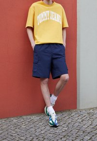 Žluté bavlněné tričko s krátkým rukávem s nápisem "TOMMY JEANS" v bílé barvě, kombinované s námořnickými cargo šortkami a vícebarevnými teniskami. Texturované pozadí.