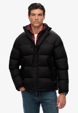 Chaqueta de plumas negra con diseño acolchado, cierre delantero con cremallera, bolsillos laterales y cuello alto, superpuesta sobre una camisa de cuadros roja. Jeans de mezclilla visibles.