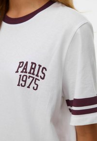 Balts kokvilnas T-krekls ar bordo ribotas apkakles, īsām piedurknēm un tekstu "PARĪZE 1975" bordo krāsā uz krūšu.