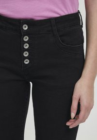 Jeans negros de cintura alta con cierre de cinco botones, que cuentan con bolsillos delanteros y detalles de costura sutiles, fabricados en tela de mezclilla.