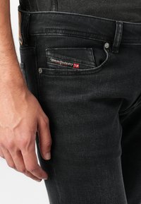 Jeans negre din denim cu o croială slim, având un eticheta vizibilă a brandului pe buzunarul din spate, nasture metalic și țesătură texturată.