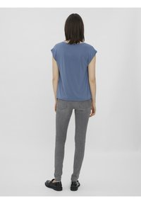 Femme portant un haut bleu à manches courtes, un jean skinny gris et des chaussures noires, debout de dos devant un fond clair uni.