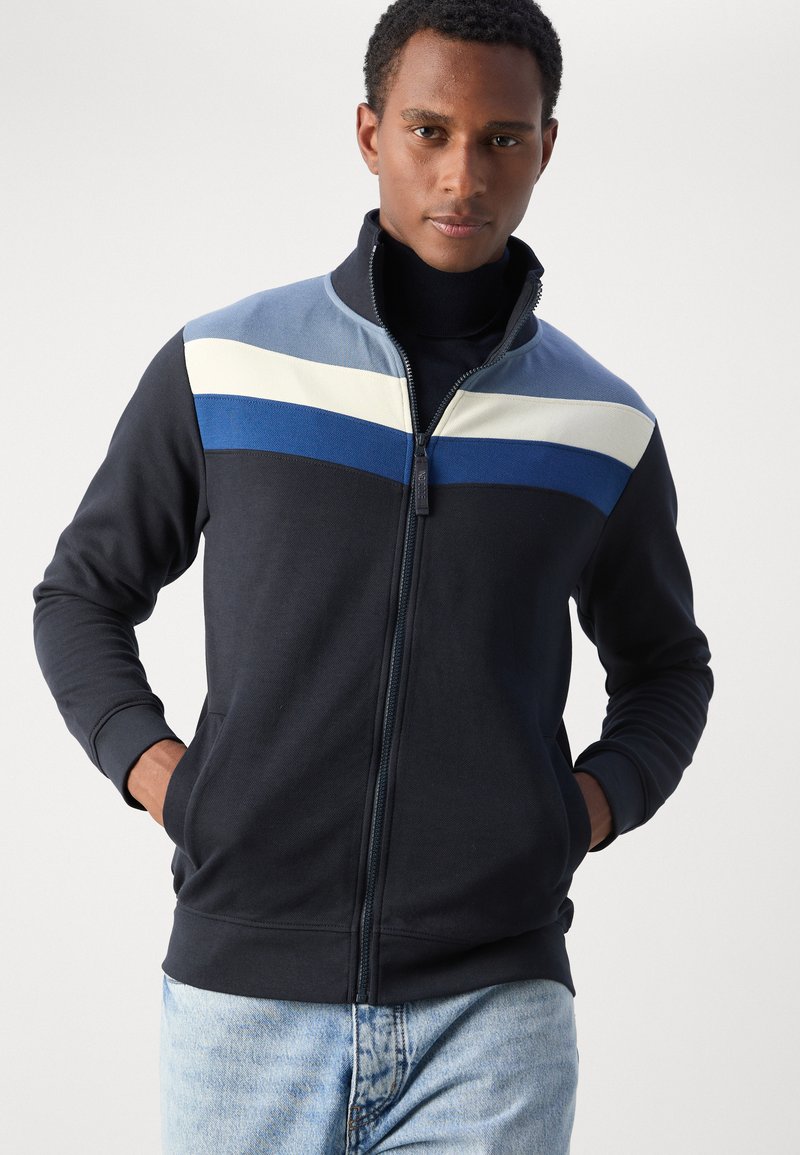 Veste zippée en tissu jersey, avec un bloc de couleurs dans les tons navy, bleu clair, blanc et bleu royal. Manches longues avec des poignets côtelés.