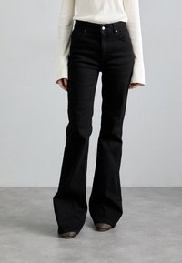 Black flared jeans with a high waist, crafted from denim. Features front pockets and a classic button closure. Worn with a white ribbed top.