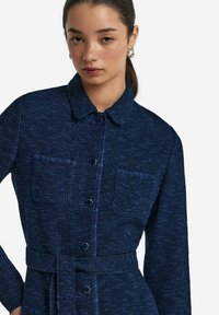 Marinblå texturerad knapp-up klänning med krage, två bröstfickor och matchande midjebälte. Slät tyg med subtila variationer.