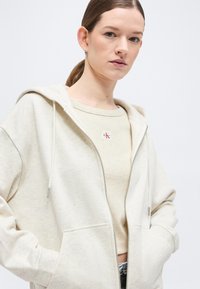 Νεαρή γυναίκα με ανοιχτό καστανά μαλλιά σε αλογοουρά, φορώντας εκρού φούτερ με φερμουάρ και μπεζ πουλόβερ Calvin Klein, με τα χέρια στις τσέπες.