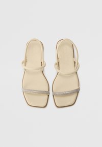 Sandales plates beiges avec deux fines lanières et une bande décorative en strass sur le devant du pied. Fini lisse et mat, design à bout carré.