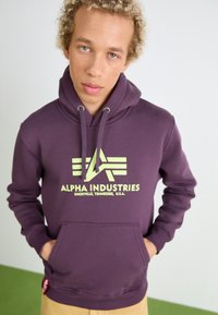 Sweat à capuche violet avec une poche avant, cordon de serrage à la capuche, et un logo jaune avec du texte stylisé et des rayures. Texture de tissu douce visible.