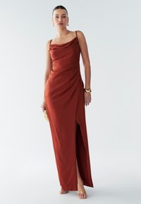 Maxiklänning i djup rostfärg med draperad halsringning, sidospalt och veckad detaljering. Spaghettiband och slät tygfinish. Guldaccessoarer.