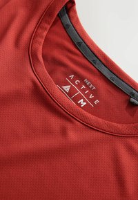 Rotes Sportoberteil aus strukturiertem Stoff mit rundem Ausschnitt und sichtbarem Branding im Kragen.