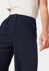 Kobieta w granatowych spodniach z wysokim stanem, z plisami i guzikiem, do których założona jest włożona do środka biała bluzka z krótkim rękawem.