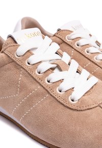 Zapatillas de ante color tierra con cordones blancos, ojales redondos y suela texturizada. Marca dorada en la lengüeta y acentos de costura blanca.