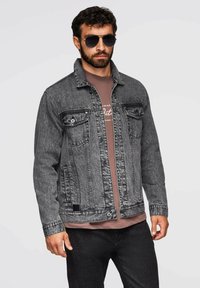 Giacca di jeans grigia con una finitura sbiadita. Presenta una classica chiusura a bottoni frontale, due tasche sul petto e un colletto standard. Indossata sopra una t-shirt marrone.