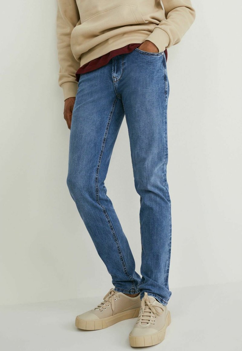 Jean en denim bleu avec une coupe slim, présentant un léger effet de froissement et un design classique à cinq poches, associé à des baskets beige clair.