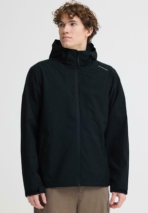 Noormees, kes kannab musta tõmblukuga kapuutsiga jopet, millel on väikene logo rinnal, seisab ühetaolise hele taustaga ees.