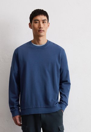 Sweatshirt en coton bleu à col ras du cou avec manches longues, poignets et ourlet côtelés, porté par-dessus un sous-vêtement gris, associé avec un pantalon sombre.