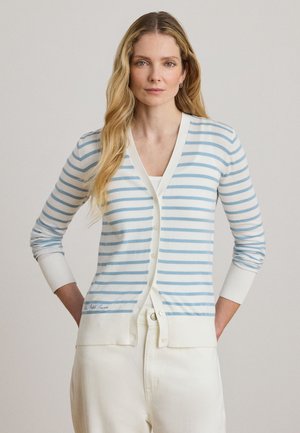 Femme aux longs cheveux blonds portant un cardigan à rayures blanches et bleu clair ainsi qu'un pantalon blanc, debout les mains dans les poches devant un fond uni.
