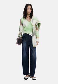 Pull en tricot vert à motifs géométriques, associé à un pantalon en denim foncé ample. Le mannequin tient un petit sac et porte des ballerines noires et blanches.