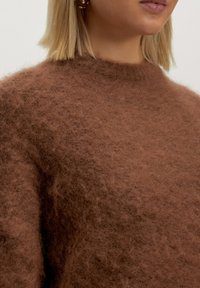 Nahaufnahme einer Person mit einem flauschigen braunen Pullover und goldenen Steckerohrringen, die schulterlanges blondes Haar und einen neutralen Lipgloss trägt.