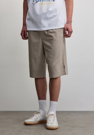 Homme portant un short beige jusqu'aux genoux, des chaussettes blanches, des baskets blanches et un t-shirt blanc avec un motif graphique, debout sur un sol gris contre un mur uni.
