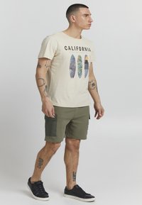 T-shirt en coton beige avec un graphique "California" et un design de feuille de palmier. Shorts vert olive avec poches latérales. Baskets noires.