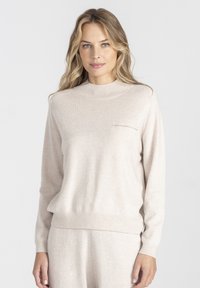 Jasno beżowy sweter z dzianiny o wysokim kołnierzu i długich rękawach. Posiada małą kieszonkę na piersi oraz ściągacze na mankietach i dolnej krawędzi. Miękka faktura.