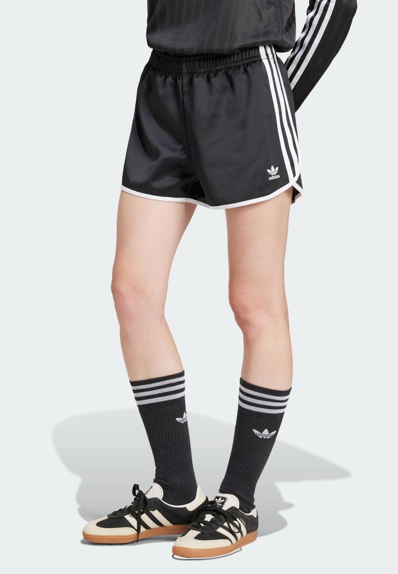 Person som bär svarta Adidas träningsshorts med vita ränder, svarta Adidas strumpor med hög krage, och svart-vita Adidas sneakers, stående mot en ljus bakgrund.