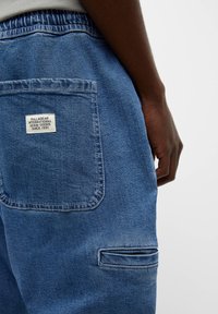 Close-up van de achterzak op een blauwere denim broek met een stoffen label waarop staat "Pull&Bear International Denim Goods Since 1991" en een hand van een persoon in de buurt.