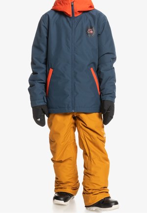 Blaue Schneejacke mit orangefarbenen Akzenten, Reißverschlussfront und Logo. Kombiniert mit orangen Schneehosen, schwarzen Handschuhen und schwarzen Stiefeln für kaltes Wetter.
