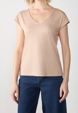 Femme portant un haut beige à manches courtes en V et un jean bleu, se tenant devant un fond clair uni.