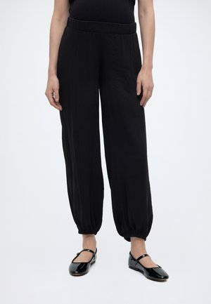 Femme portant un pantalon noir ample avec des poignets élastiques et des chaussures Mary Jane en cuir verni noir, debout devant un fond blanc uni.