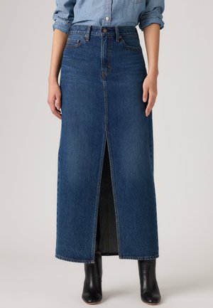 Personne portant une jupe midi en denim bleu avec une fente à l'avant, associée à une chemise en denim rentrée et des bottines noires.