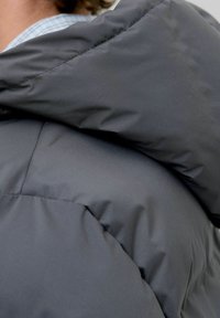 Close-up of a person wearing a dark gray padded jacket with a large collar, partly showing a blue checkered shirt underneath.